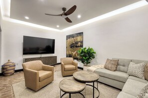 Ein modernes Wohnzimmer mit Flachbildfernseher, zwei beigen Sesseln, einem grauen Sofa und einem Deckenventilator.
