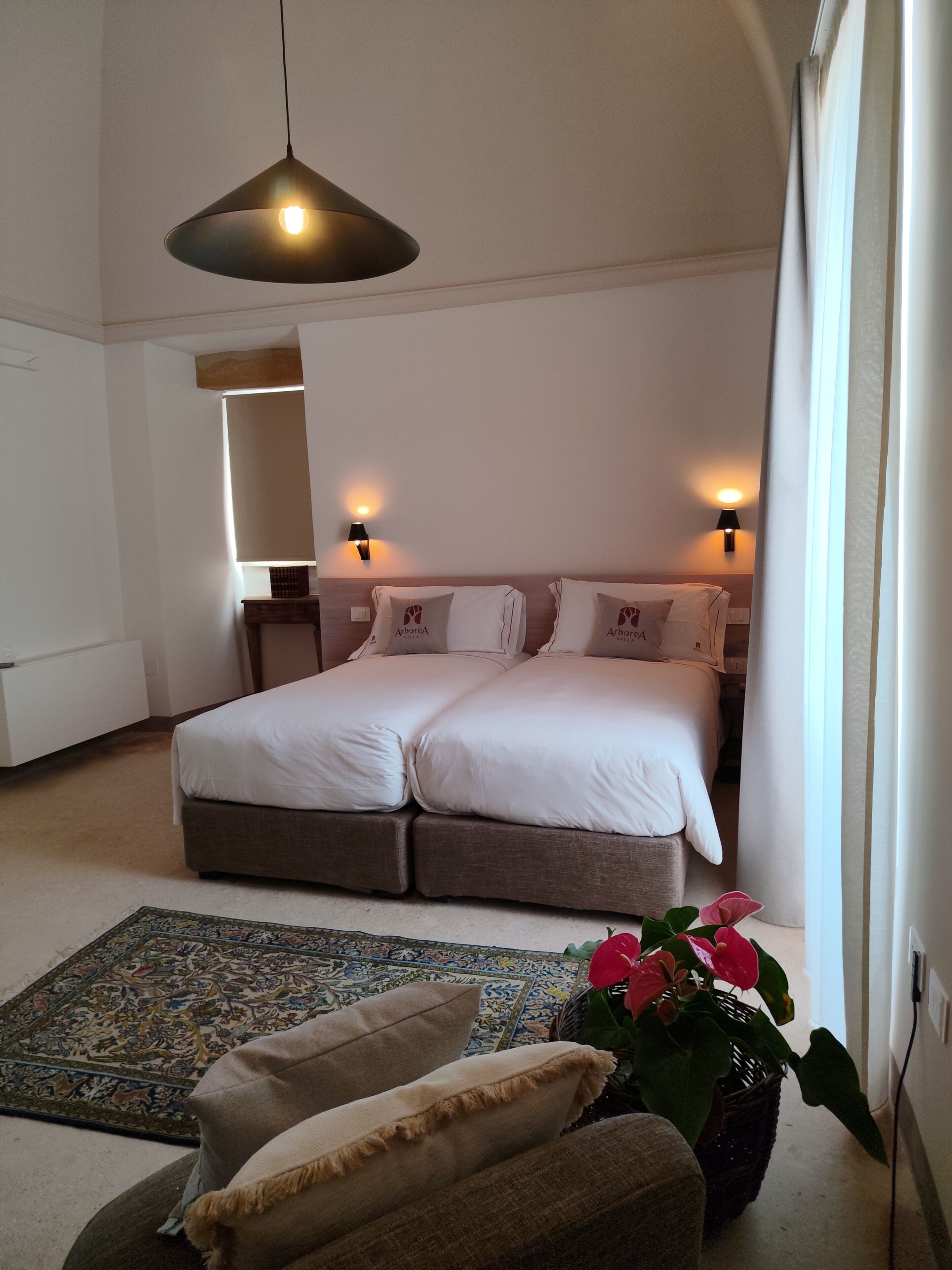 A bedroom with a double bed, bedside lamps, a hanging light fixture, a patterned rug, and a potted plant.
