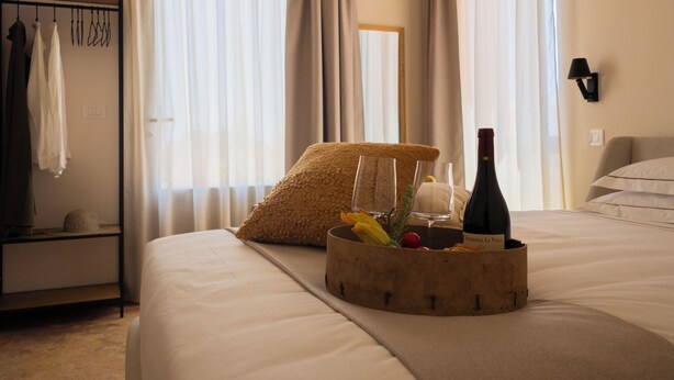 Quarto de hotel com uma cama, uma mesinha de cabeceira com uma garrafa de vinho, copos e uma cesta de frutas.