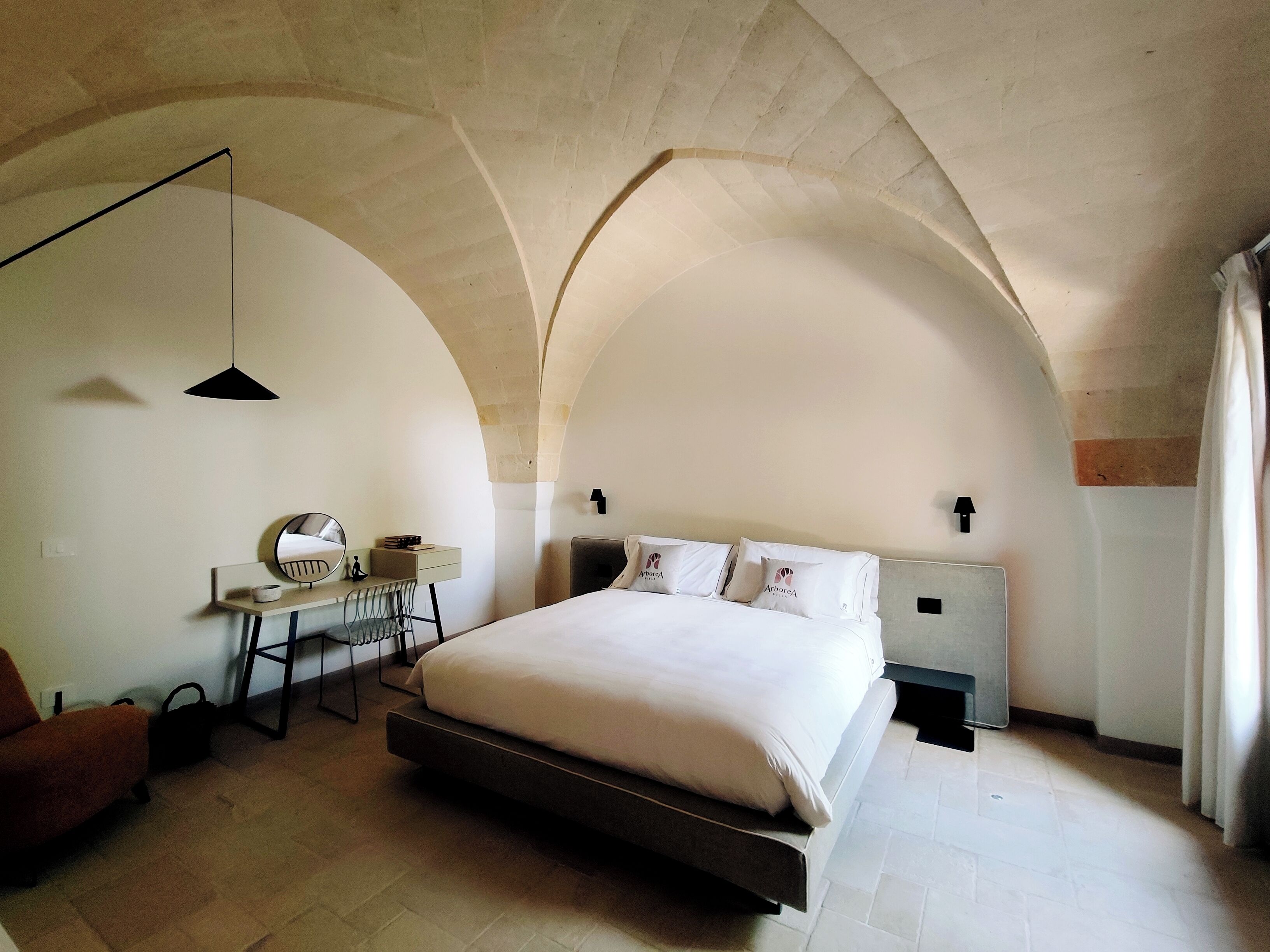 A bedroom with a large bed, a desk with a chair, a mirror, and a hanging light fixture.