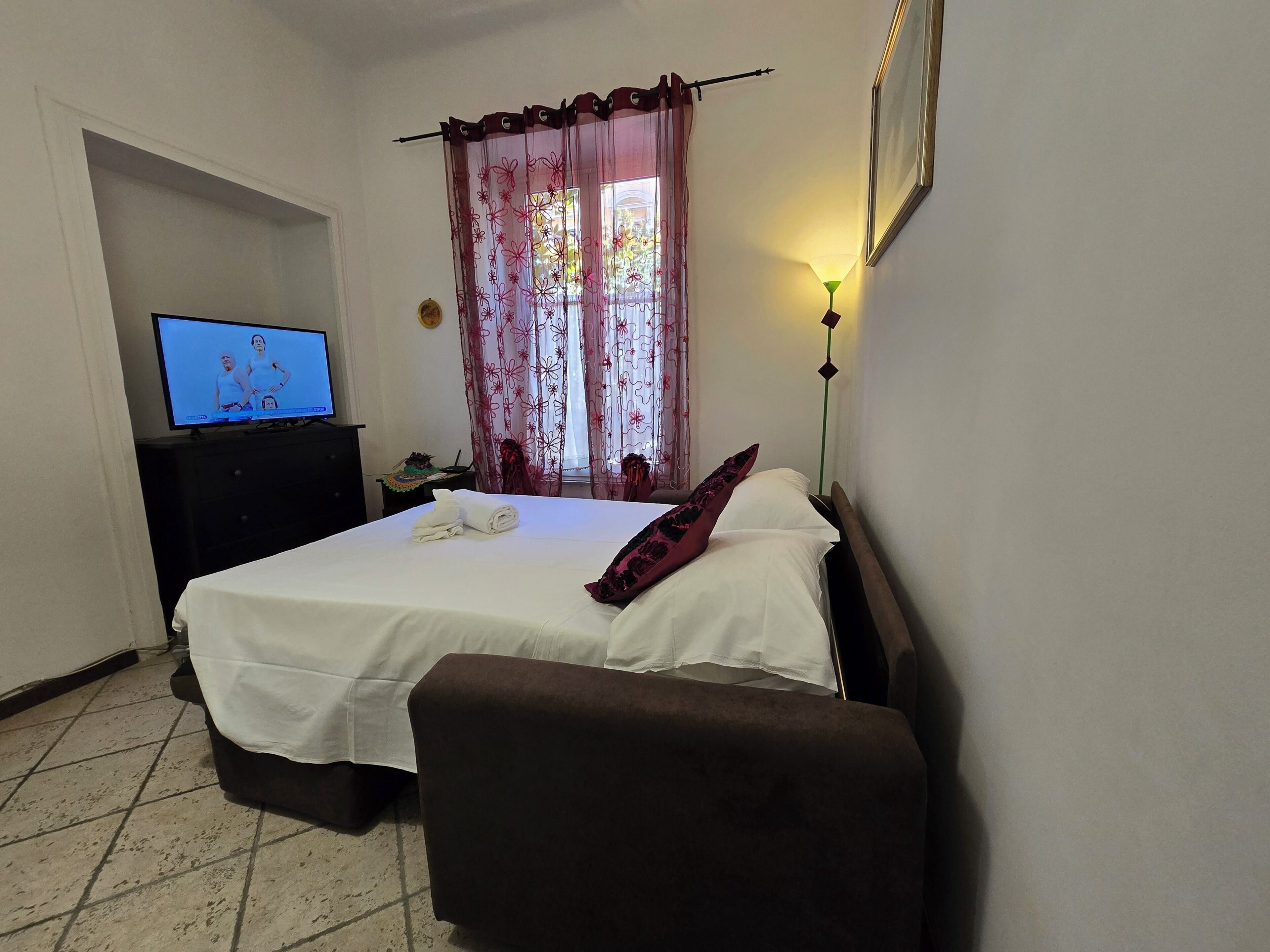 A hotel room with a bed, a TV, a nightstand, a lamp, and a window with curtains.