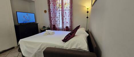 A hotel room with a bed, a TV, a nightstand, a lamp, and a window with curtains.