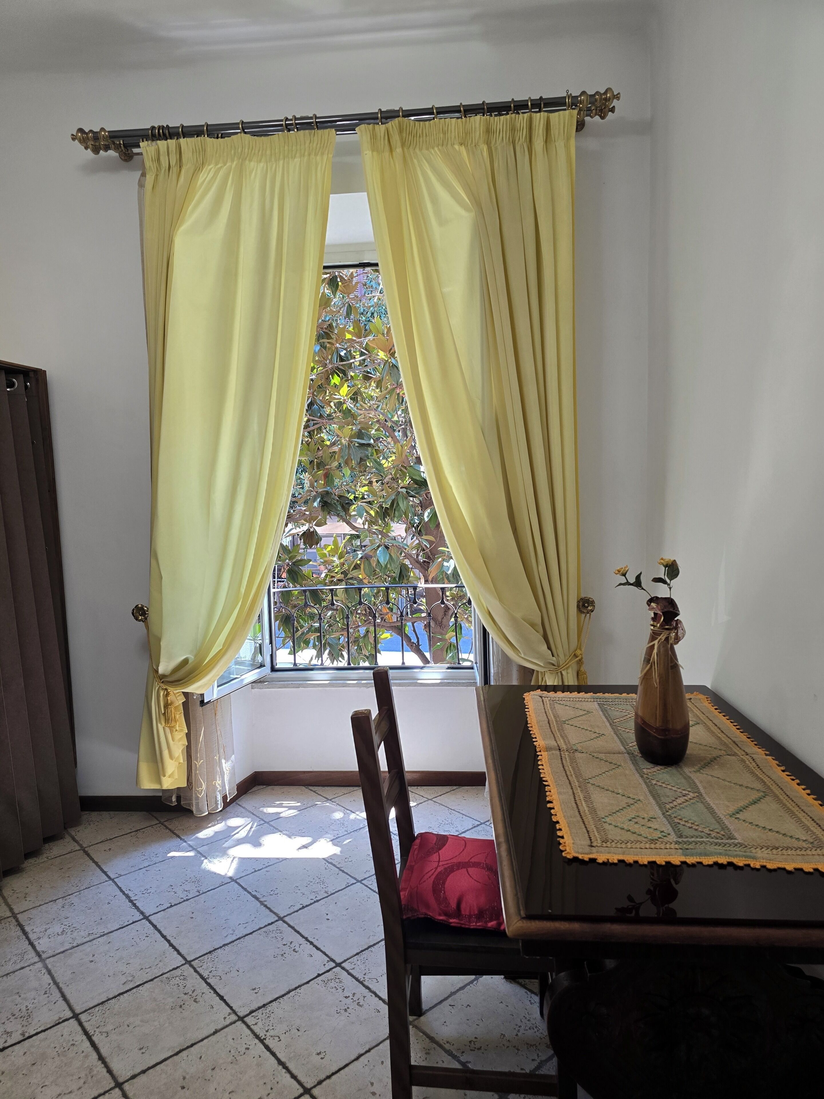 Una habitación con ventana, cortinas, una silla, una mesa y un florero con flores.