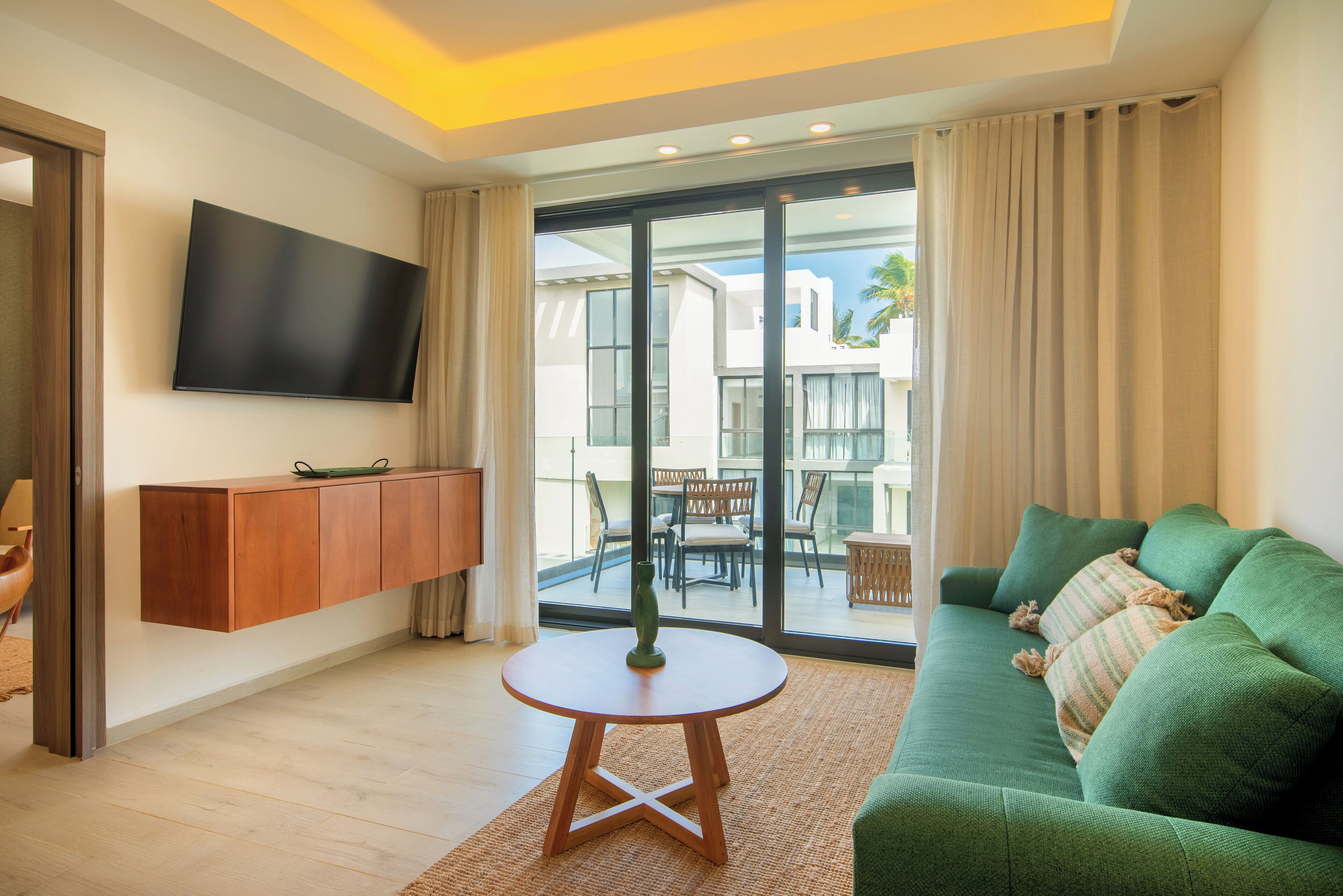 A modern living room with a flat-screen TV mounted on the wall, a green sofa, a small round table, and a view of the outside balcony.