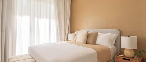 Una habitación de hotel con una cama grande, mesitas de noche, una lámpara y cortinas translúcidas.