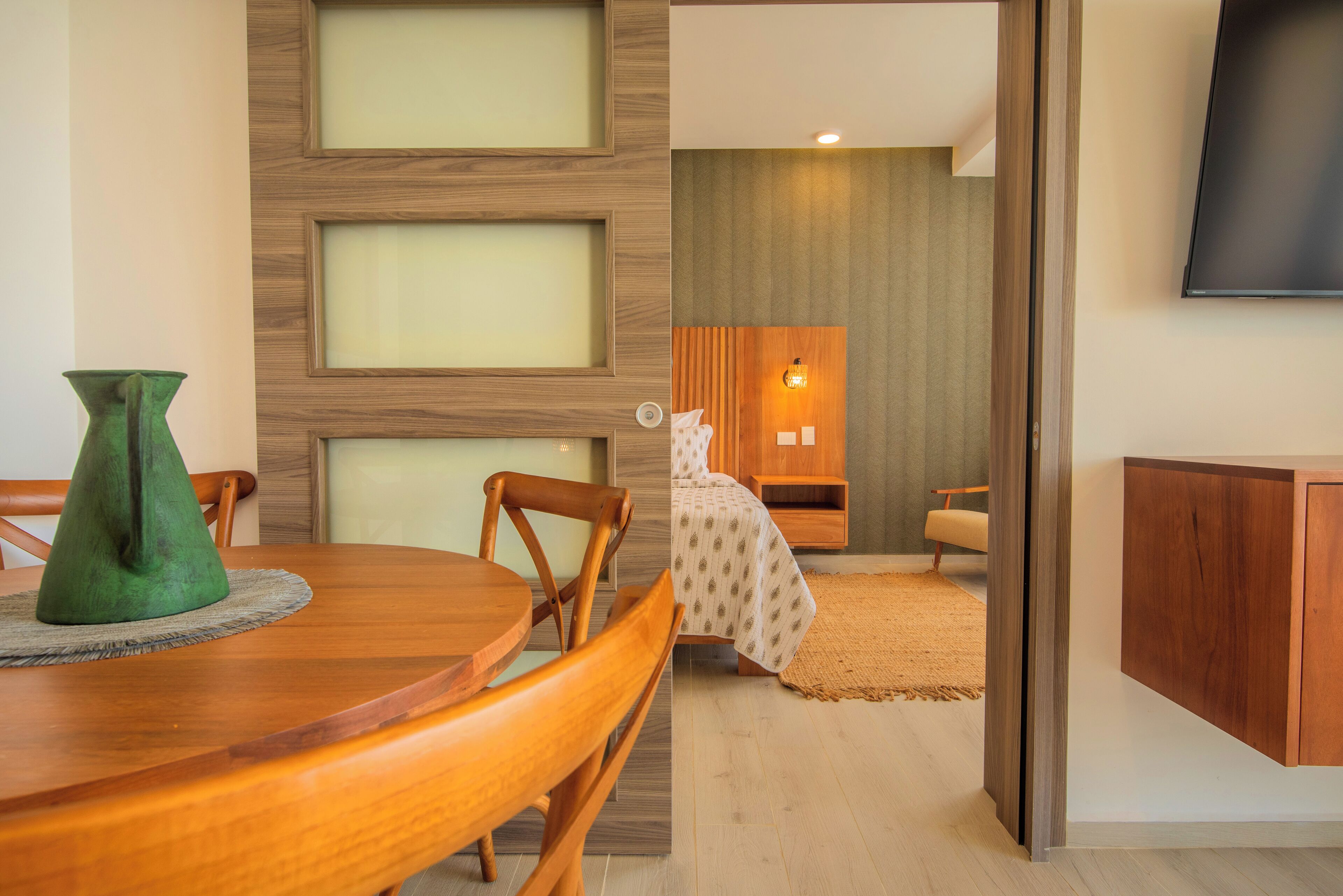 Una habitación de hotel moderna con una mesa de comedor de madera, un jarrón verde y una cama con una colcha estampada.