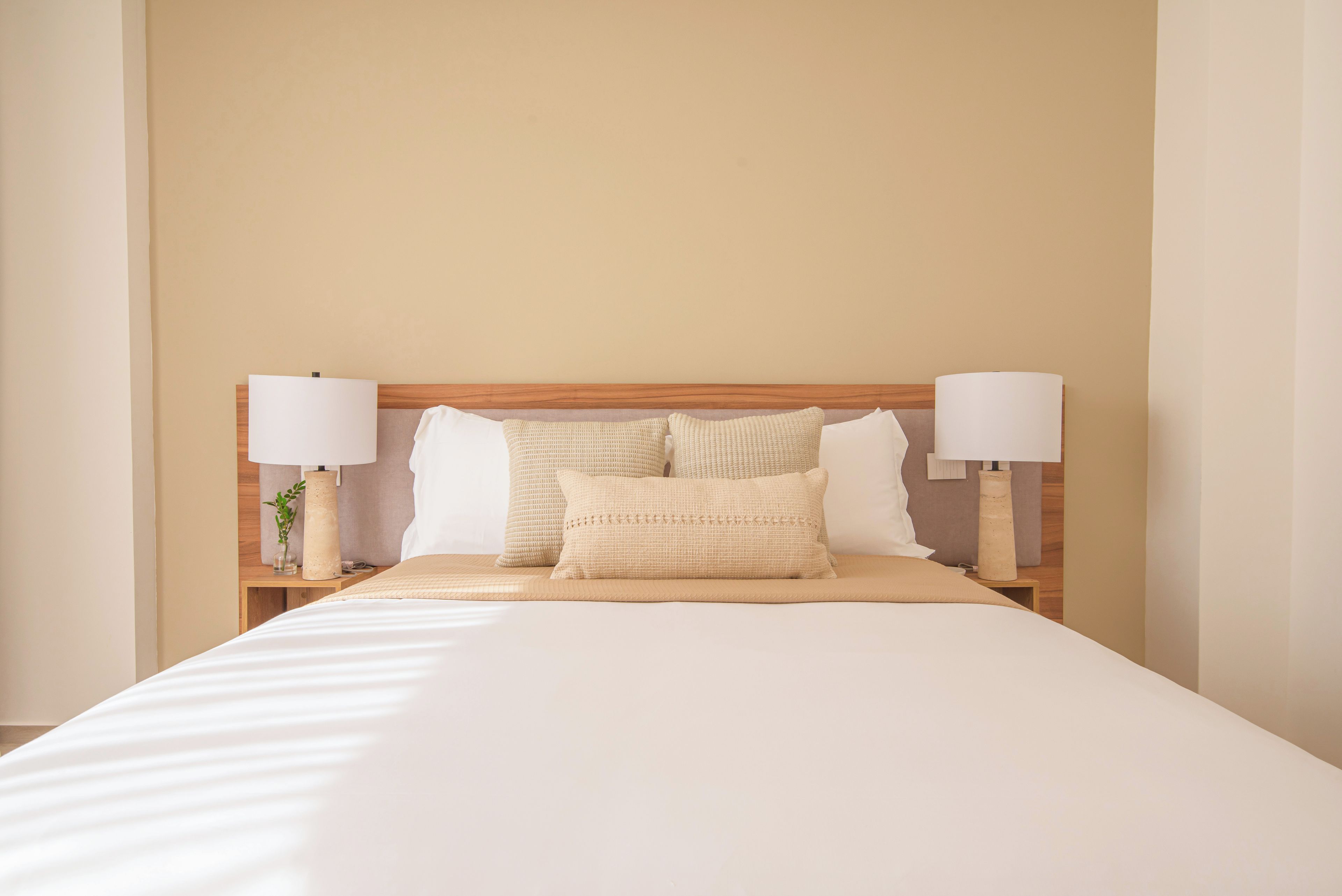 Uma cama bem arrumada com cabeceira de madeira, duas mesas de cabeceira com abajures e um vaso com uma planta em uma delas.