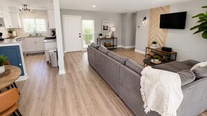 Een moderne keuken met witte kasten, een eethoek met een houten tafel, een grijze bank en een aan de muur bevestigde televisie.