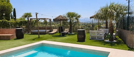 A rectangular swimming pool with a tiled edge, surrounded by a grassy area with palm trees and a wooden pergola.