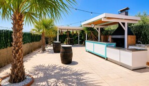 Een terras buiten met een bar, een grill en palmbomen.