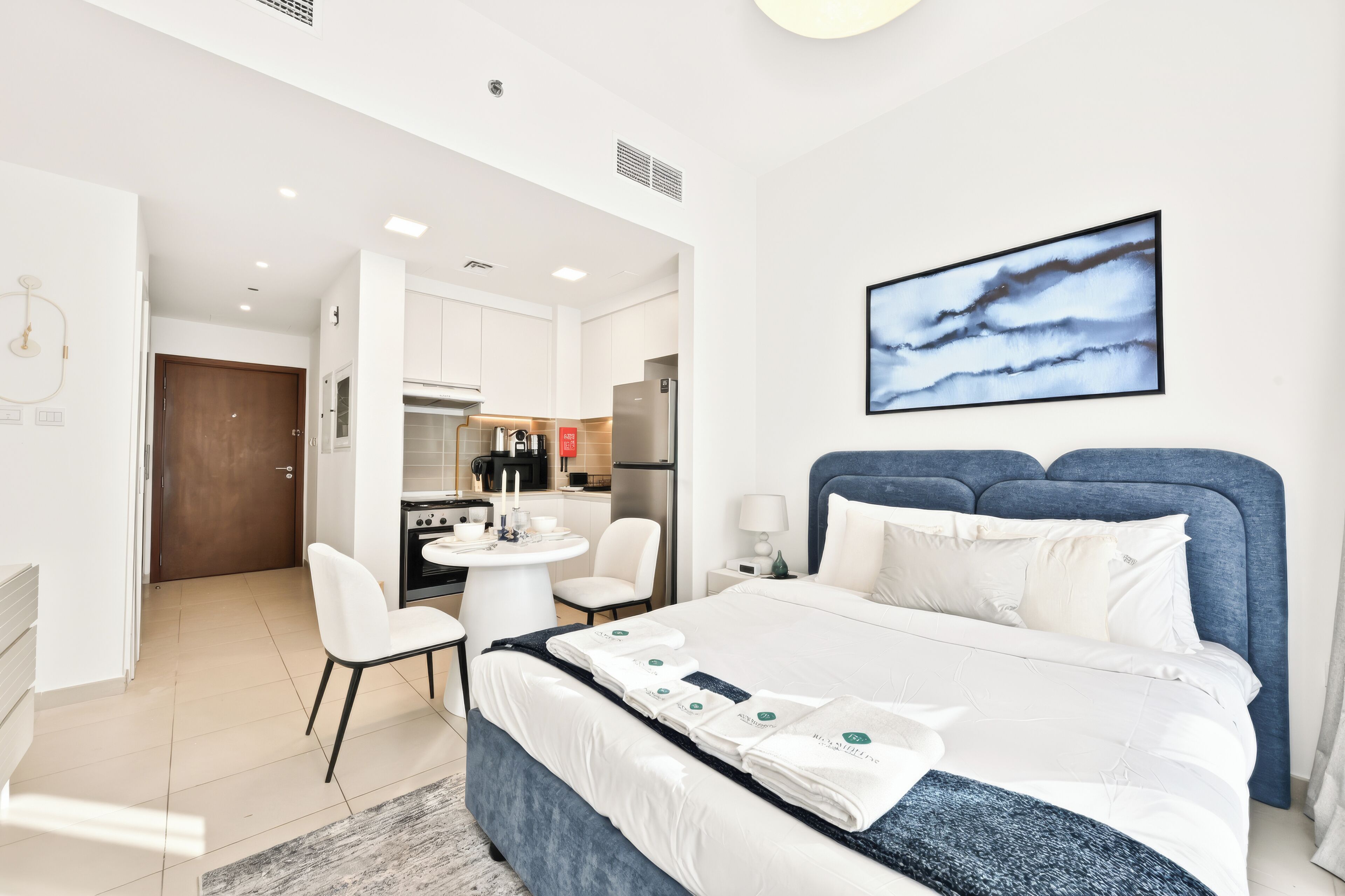 A modern hotel room with a bed, a dining table, chairs, a kitchen area, and a painting on the wall.