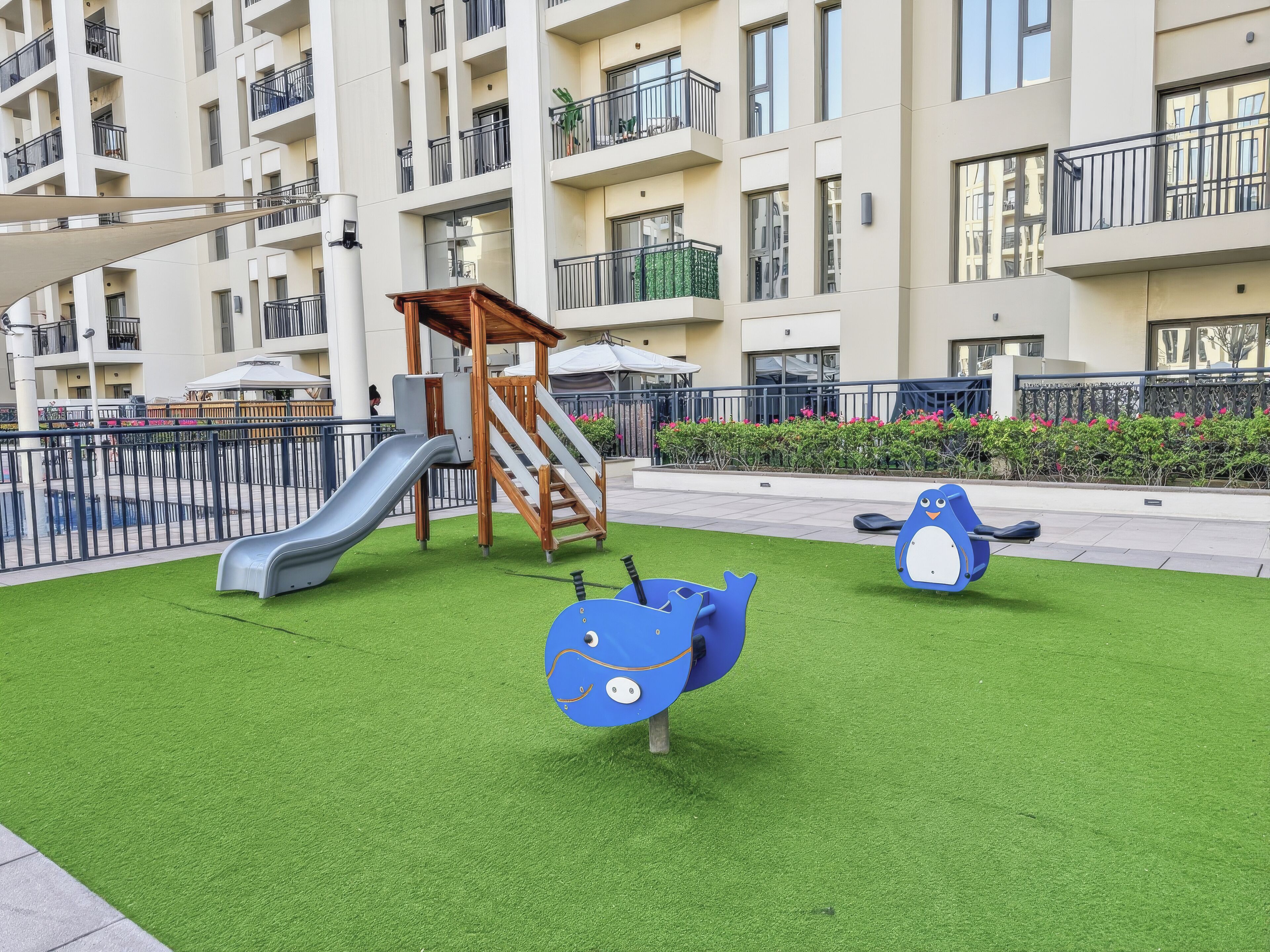 Ein Spielplatz mit Rutsche und Klettergerüst, umgeben von einem Zaun und Grünpflanzen.