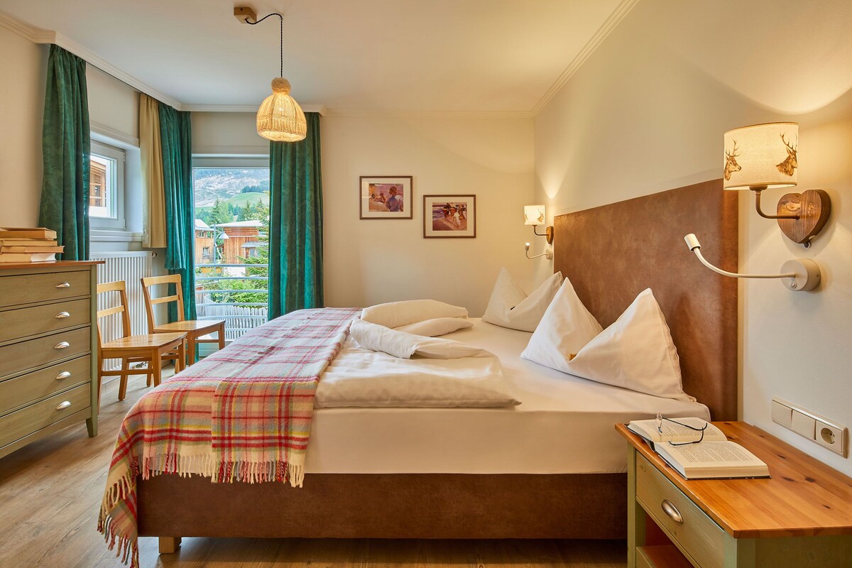 A hotel room with a bed, bedside table, dresser, and a view of the outside.