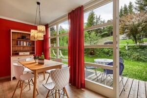 Una habitación con una mesa de madera, sillas, una estantería y un ventanal con vistas a un jardín.