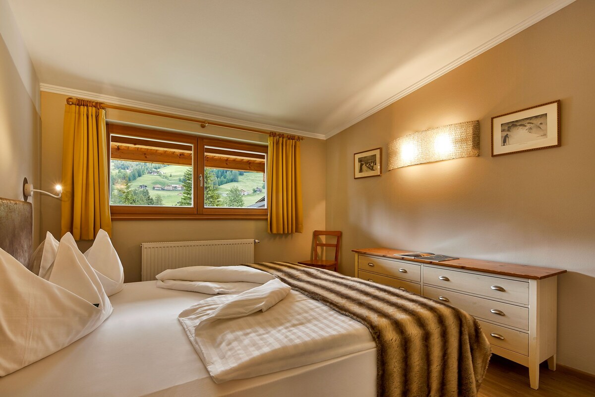 A hotel room with a bed, a wooden dresser, a chair, a window with curtains, and a wall-mounted light fixture.