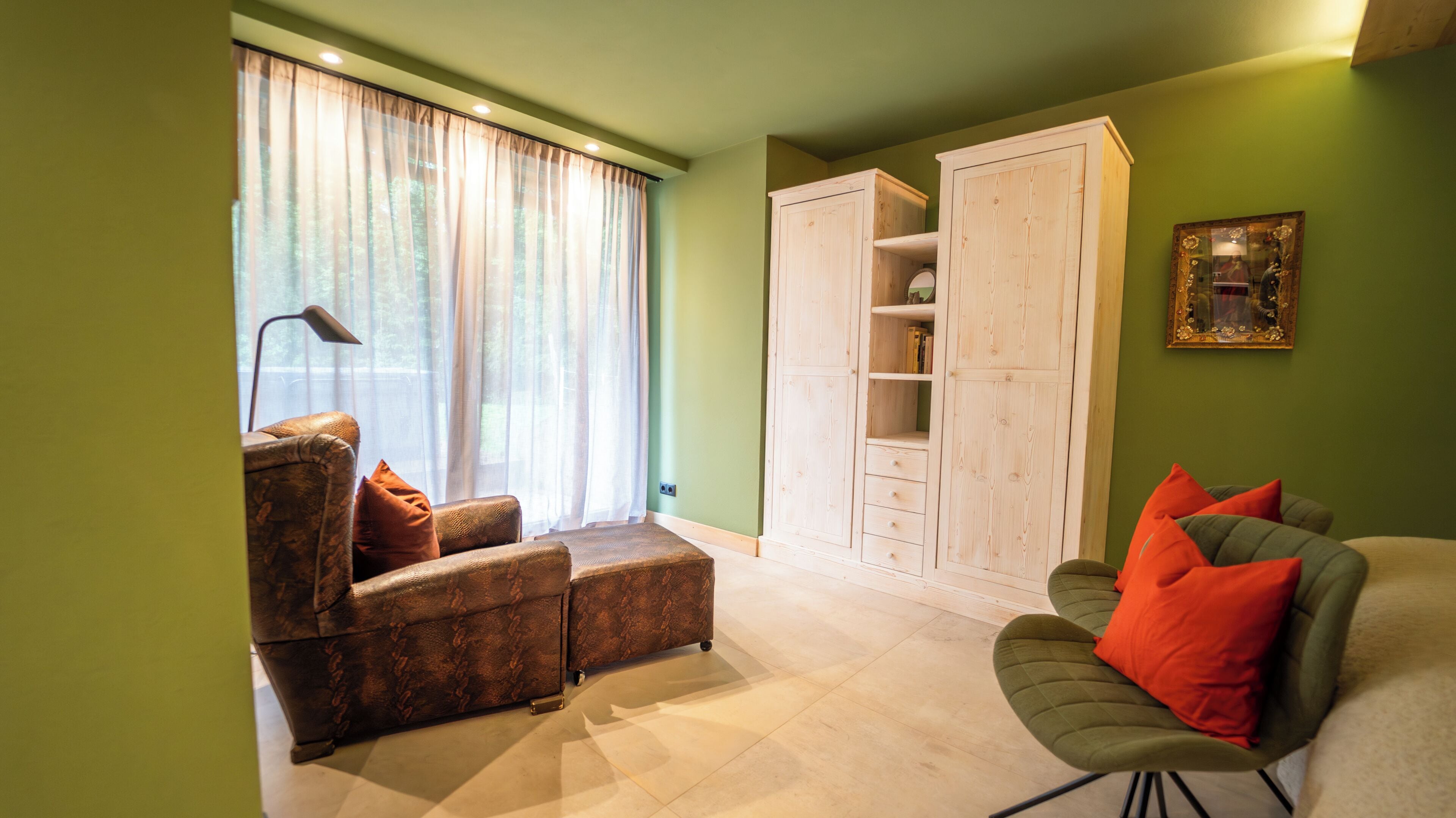 A living room with a sofa, ottoman, and a green armchair. There is a large wardrobe and a painting on the wall.