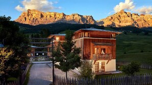 A wooden chalet with a balcony, surrounded by a wooden fence, set against a mountainous backdrop. - Appartamenti La Sajun e Les Viles (Badia)