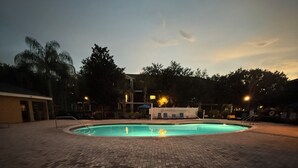 Ein Schwimmbad mit Pflasterterrasse, umgeben von Bäumen und einem Gebäude mit Beleuchtung.