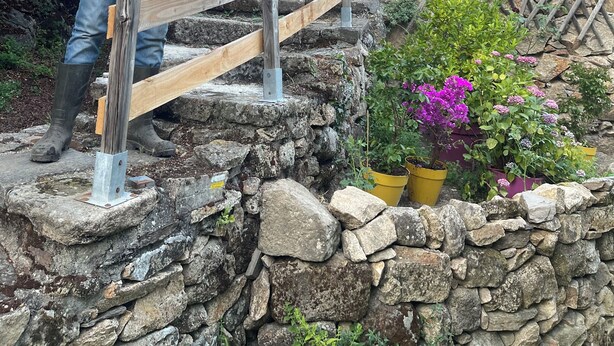Eine Person gießt Pflanzen auf einer Holztreppe mit Steinwänden und Blumentöpfen.