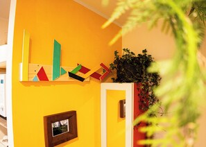 Una pared amarilla con formas geométricas, un cuadro enmarcado y una planta en maceta.