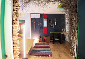 Una habitación con una estantería de madera, un tapete colorido, una guitarra y una pequeña mesa con cubierta verde.