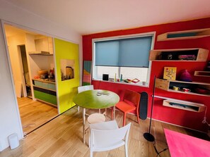 Quarto pequeno com mesa de jantar, cadeiras, janela com persianas, estante de livros e uma área de cozinha compacta.