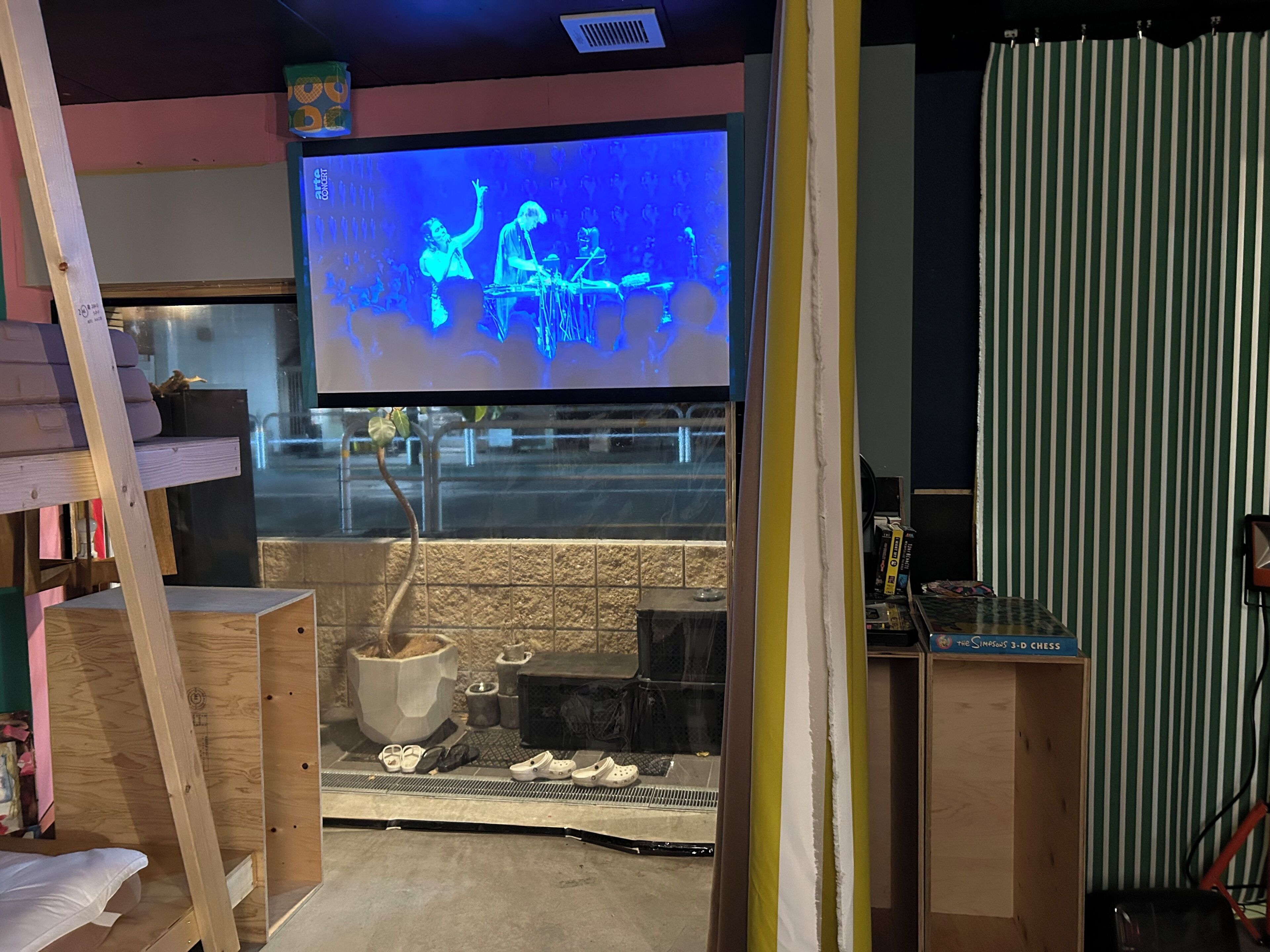 A room with a television displaying a band performance, a tiled wall, and a window with a view of the street.