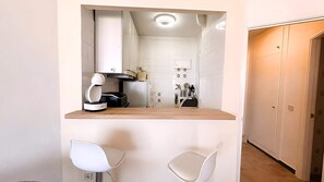 Eine kompakte Küche mit Einbauofen, Kaffeemaschine und kleiner Arbeitsfläche. Zwei weiße Stühle und eine Tür zu einem anderen Raum.
