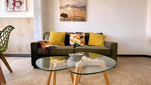Ein Wohnzimmer mit einem Glastisch, einem grauen Sofa mit gelben und bunten Kissen sowie einem gerahmten Bild eines Gebäudes an der Wand.