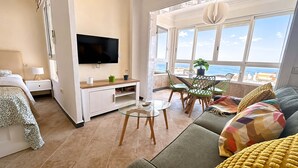 Ein modernes Wohnzimmer mit Flachbildfernseher, Essbereich mit Stühlen, einer Couch mit farbenfrohen Kissen und einem großen Fenster mit Meerblick.