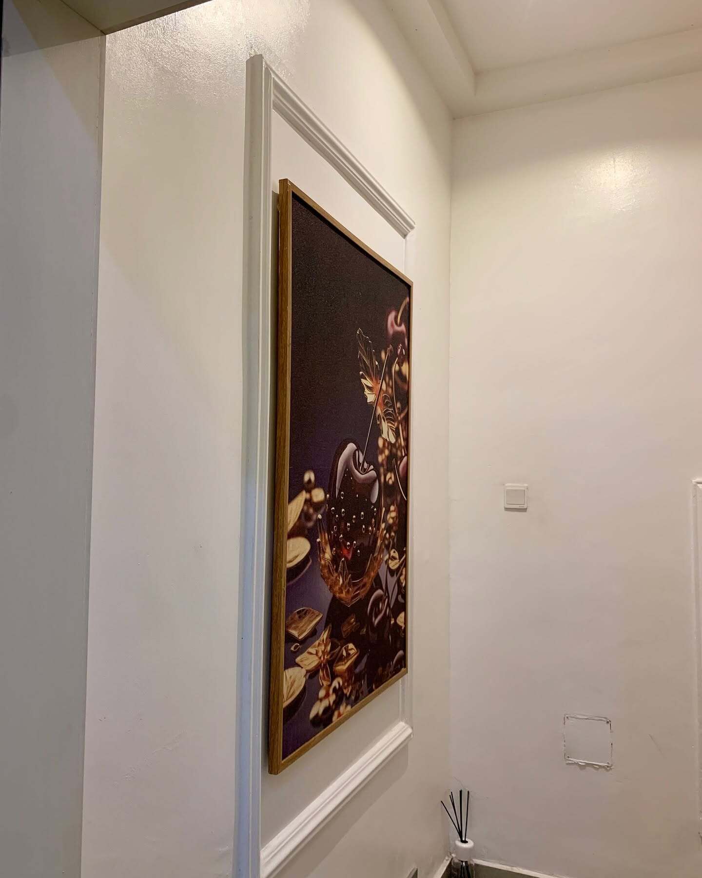 Een ingelijste afbeelding aan een witte muur in een kamer met een witte deur en een zichtbare lichtschakelaar.