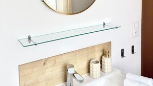 Un baño con un espejo redondo, repisa de vidrio, tocador de madera, lavamanos, toalla y una planta.