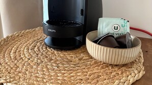 Una cafetera negra sobre un posavasos tejido, un cuenco con una cajita de cápsulas de café Dolce Gusto y un cable de alimentación rojo.