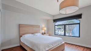 A bedroom with a wooden bed, a nightstand with a lamp, a window with a view of buildings, and a ceiling light fixture. - Lovely Beachside 2-Bedroom Apartment (Santa Monica)