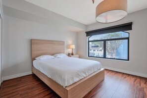 A bedroom with a wooden bed, a nightstand with a lamp, a window with a view of buildings, and a ceiling light fixture. - Lovely Beachside 2-Bedroom Apartment (Santa Monica)