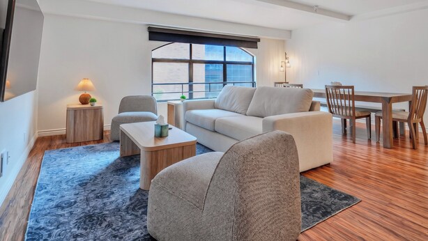 A modern living room with a sofa, armchairs, a coffee table, and a dining table with chairs. There is a large window with blinds, a lamp, and a small side table with a plant. - Lovely Beachside 2-Bedroom Apartment (Santa Monica)