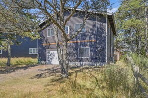 A two-story building with a garage, surrounded by trees and a wooden fence. - Large Private Deck, Game Room, Grill, Dog-Friendly (Pacific City)