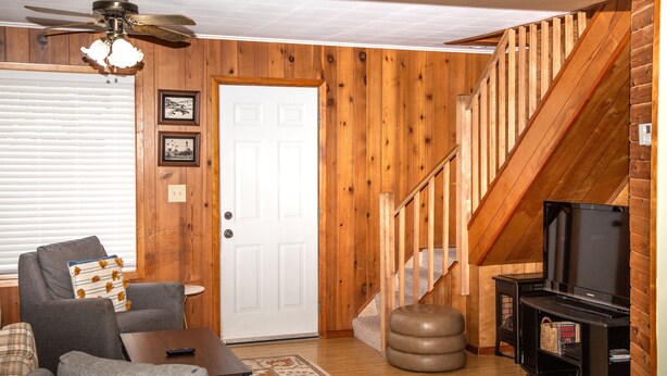 Un salon avec un ventilateur de plafond, un escalier, un téléviseur et une porte.