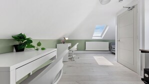 Une pièce avec un plafond en pente, un puits de lumière, un bureau blanc, une chaise, un radiateur et une plante.