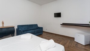 Une chambre d’hôtel moderne avec un lit, un canapé bleu, un petit réfrigérateur blanc, un téléviseur fixé au mur et un plancher en bois.