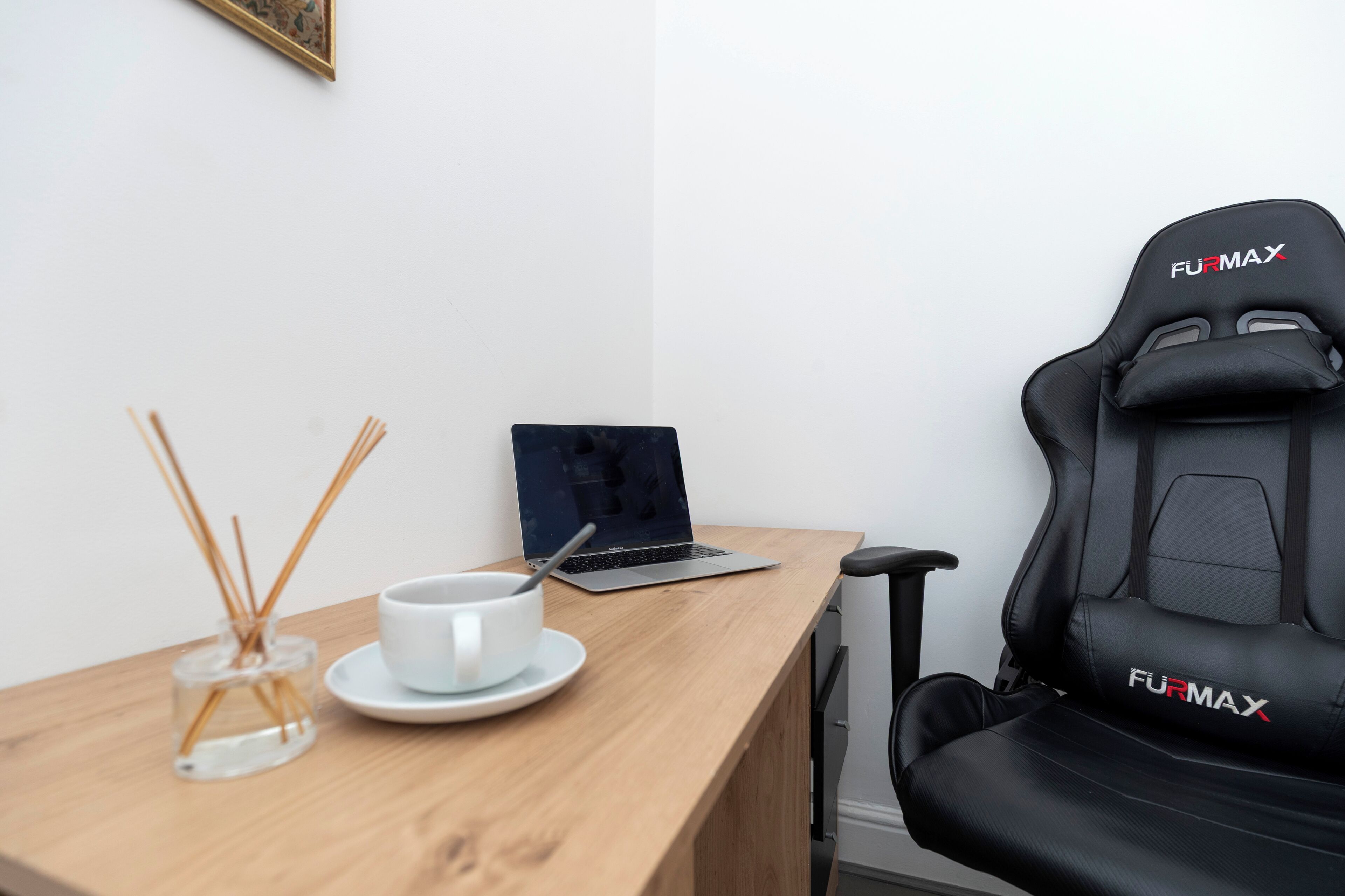 A room with a wooden desk, a laptop, a white cup with a saucer, a black gaming chair labeled 'FURMAX,' and a picture frame on the wall.