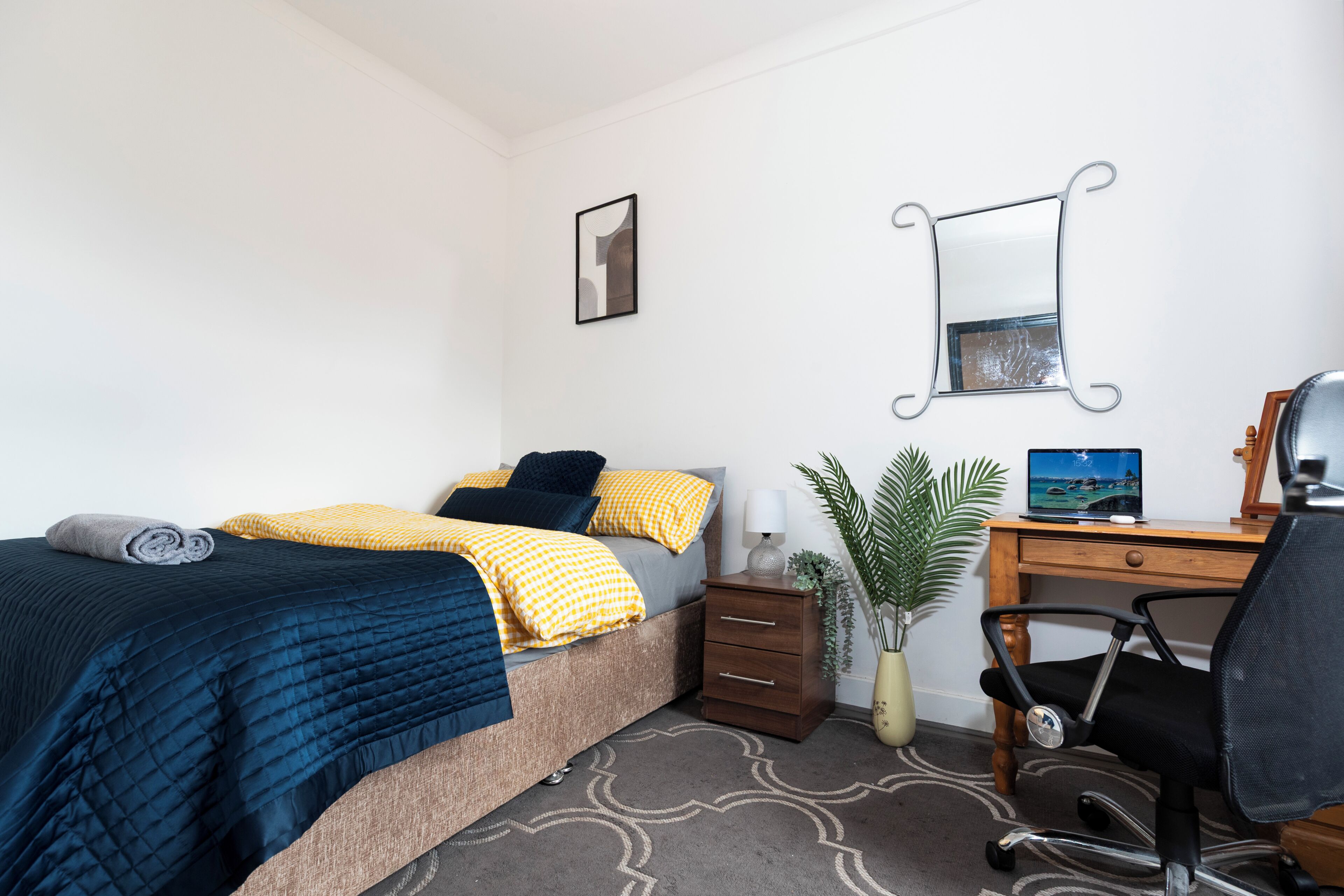 A bedroom with a bed, bedside table, lamp, chair, desk, computer, and a potted plant.