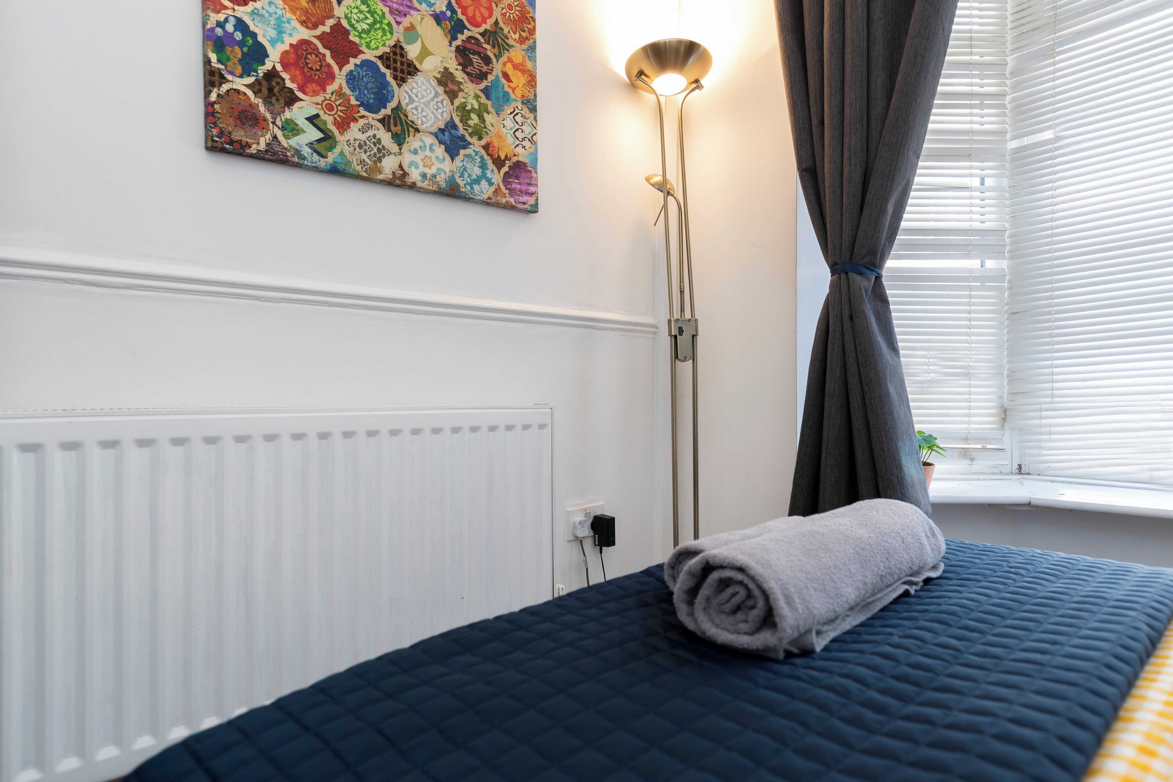 A neatly made bed with a rolled towel, a radiator, a wall-mounted light, and a window with curtains.