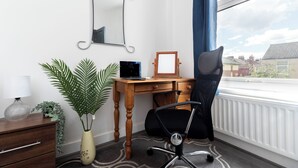 Une pièce avec un bureau en bois, un fauteuil de bureau noir, un miroir, une plante en pot et une fenêtre donnant sur des maisons.