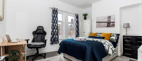 A bedroom with a bed, a desk, a chair, a dresser, a window with curtains, and a wall-mounted plant.