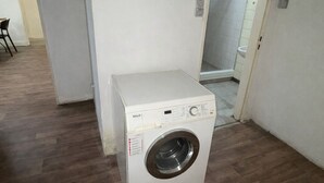 A small laundry room with a white washing machine, wooden flooring, and a door leading to a shower area.