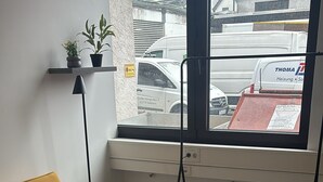 A room with a yellow sofa, a grey rug, a radiator, and a window with a view of parked cars and buildings.