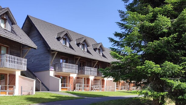 A row of wooden buildings with balconies, a paved walkway, and a tree. - A Nature Getaway in the Mountains: A Quiet Apartment (Besse-et-Saint-Anastaise)