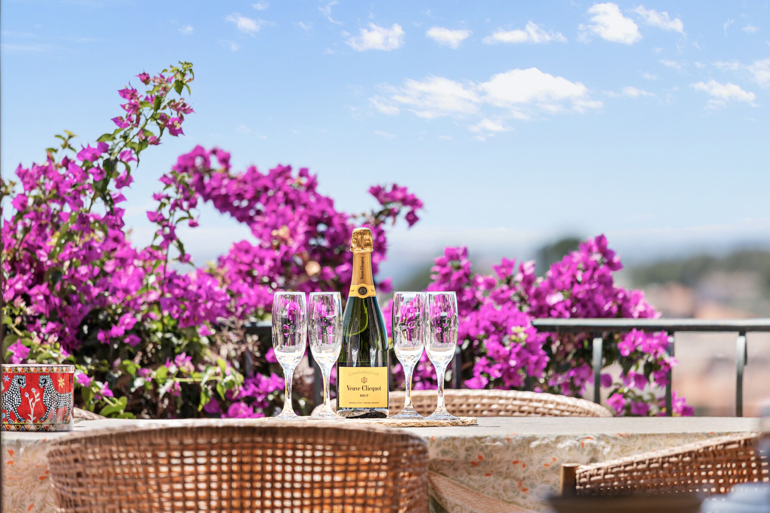 Una ampolla de champany Veuve Clicquot, quatre copes i una tassa decorativa sobre una taula de vimbles, amb vista a flors morades i un cel serè.