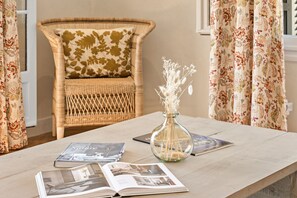 A wicker chair with a floral cushion, a glass vase with dried flowers, and open magazines on a wooden table. - Mas des Cigales – Pool & Village View (Vence)